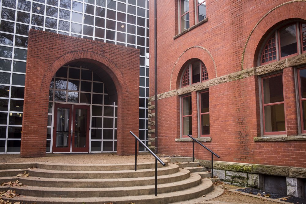 Students walking near the entrance of a brick building with large windows. One student with a backpack is walking up the stairs, while three others, dressed in business attire, are walking down, carrying boxed items. The area is surrounded by greenery.